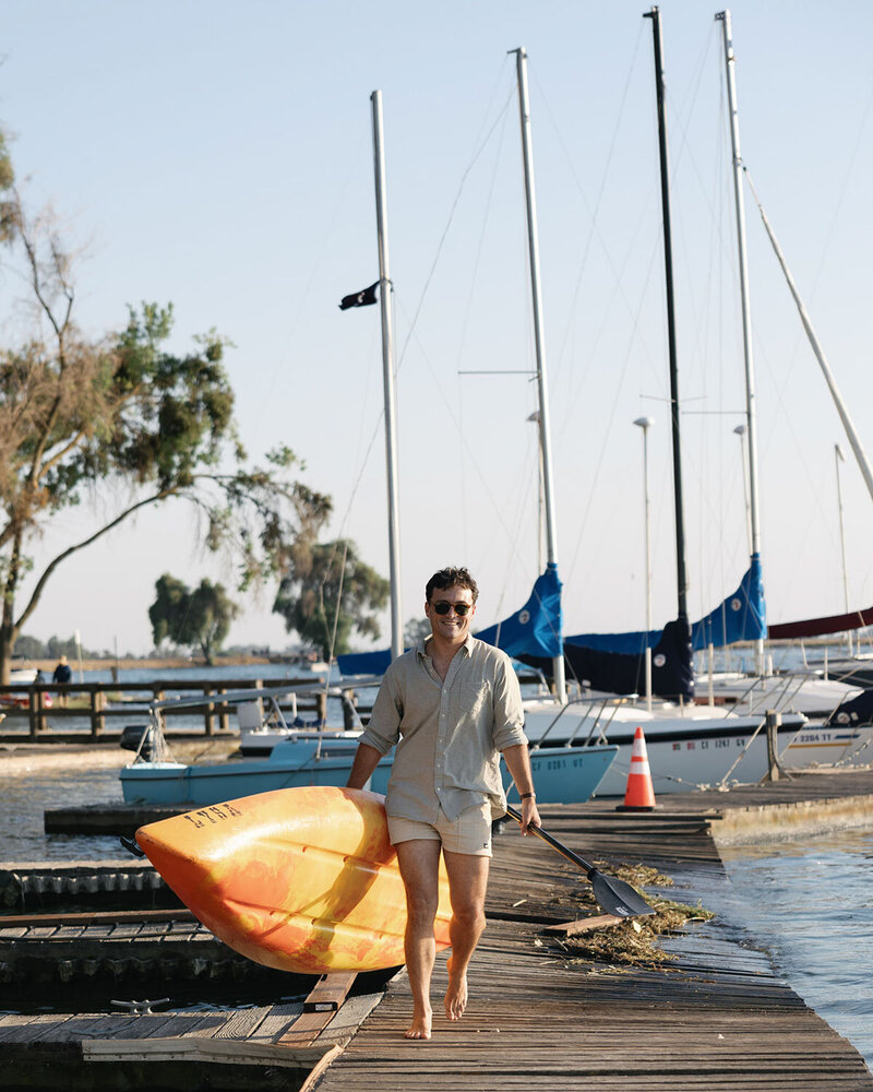 A person walks barefoot on a wooden dock, carrying a yellow kayak and a paddle.
