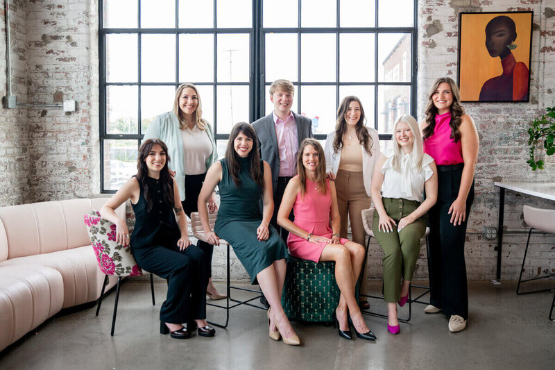 Group photo of the Pierce Public Relations team smiling in a bright, loft-style office with exposed brick walls and large windows.