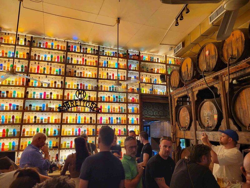 Brettos Bar in Athens, Greece, with shelves lined with colorful bottles glowing under warm lighting, wooden barrels stacked against the wall, and patrons gathered at the counter ©Stephanie Dosch | theViatrix Personalized Greece Travel Planning