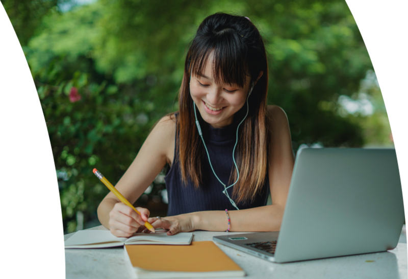 Woman sitting on a couch using a laptop, smiling as she works in a bright room with plants in the background.