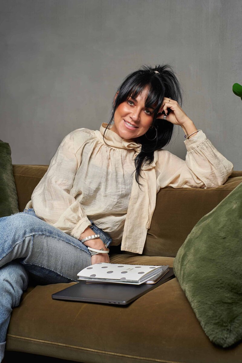 A woman with dark hair sits on a brown sofa, smiling, with her elbow resting on the armrest. An open notebook and a tablet, essential tools for custom brand and Showit web design for photographers, are on the sofa cushion next to her.