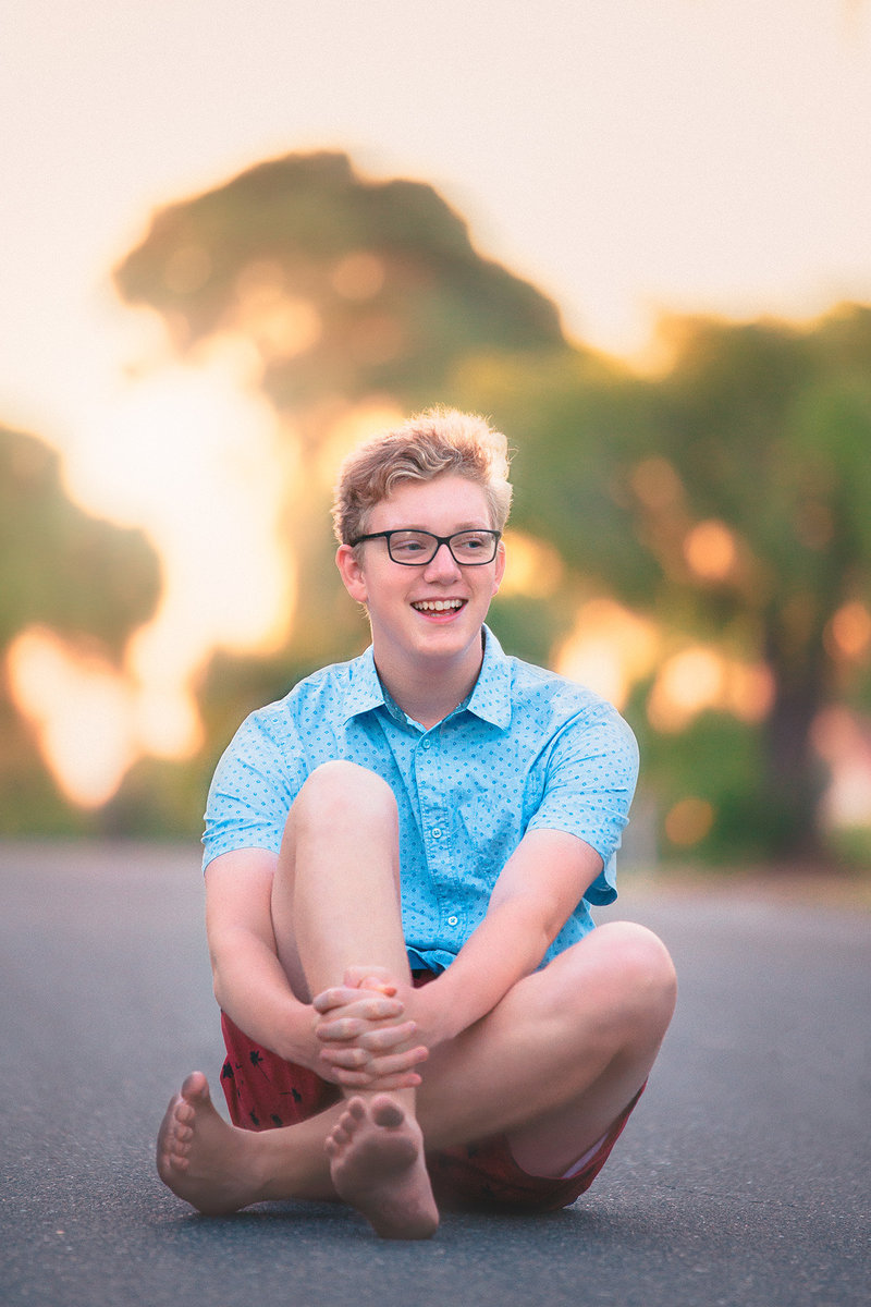 Senior picture photographer captures guy senior picture outfit ideas with a young man sitting in the road wearing a bright blue button down short sleeve shirt and red shorts