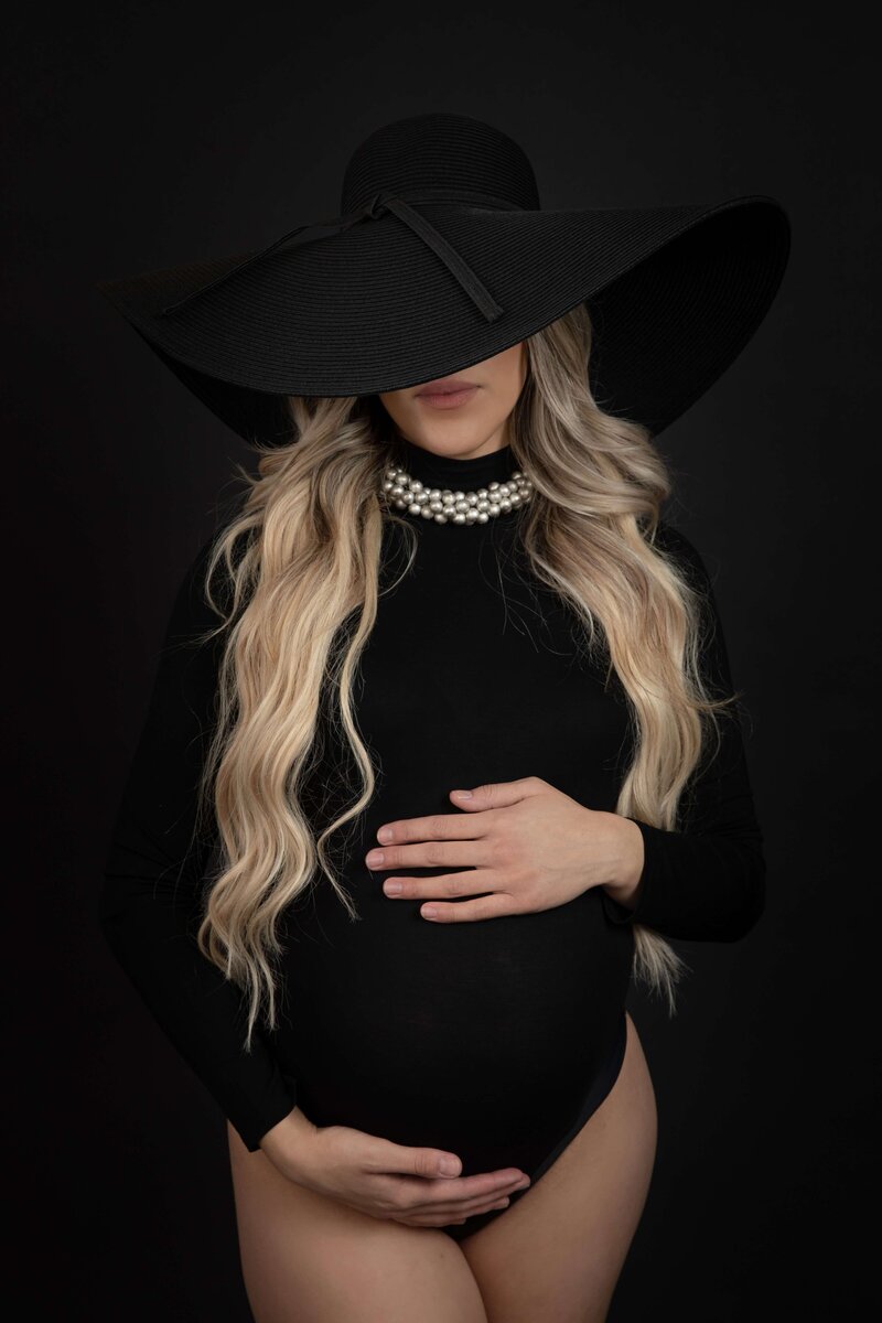 A pregnant woman wearing a black wide-brimmed hat and a strand of pearl necklace stands against a dark background. She holds her belly with both hands, partially obscuring her face with the hat. She has long, wavy blonde hair and is dressed in a black outfit.