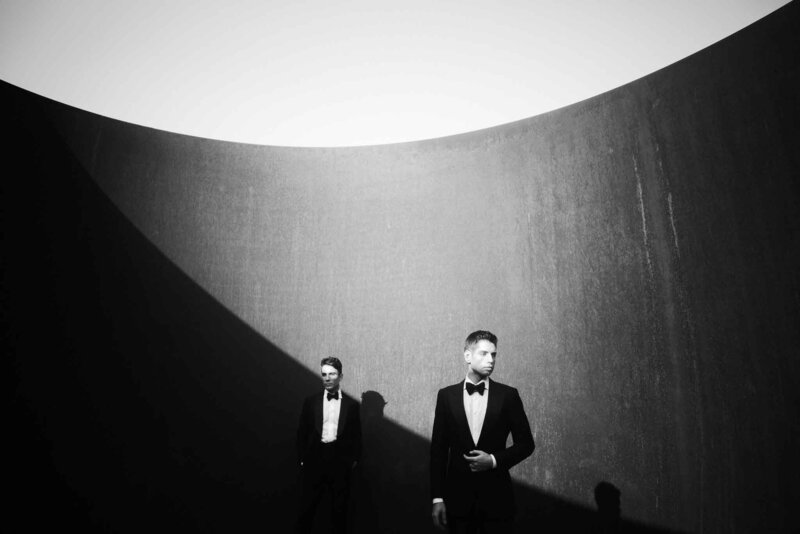 Two grooms in black tuxedos stand apart in high contrast architectural portrait