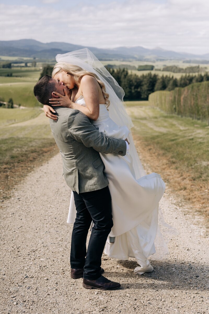 groom and bride kiss on gravel road overlooking green fields in gore new zealand
