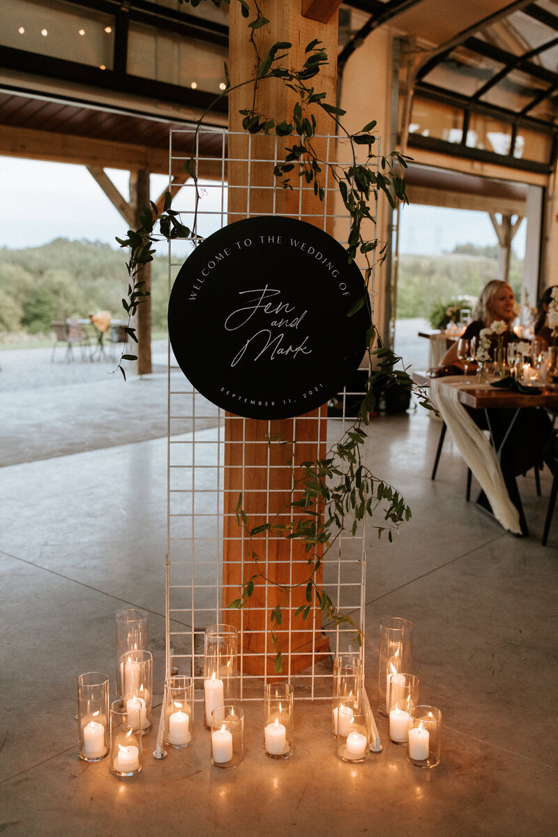 A round black modern welcome sign  is mounted on a white grid wall with greenery vines in an industrial barn with candles at the base sitting on the concrete floor at a wedding at Farmhill in Peterborough, designed by Frid Events.