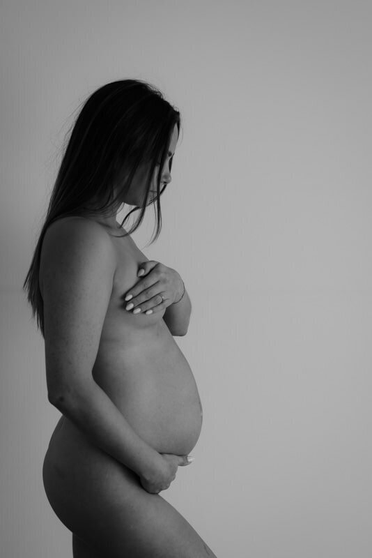 Pure foto van vrouw met zwangere buik in de studio, zwart-wit zwangerschapsfoto met zachte lichtval en minimalistische stijl.

