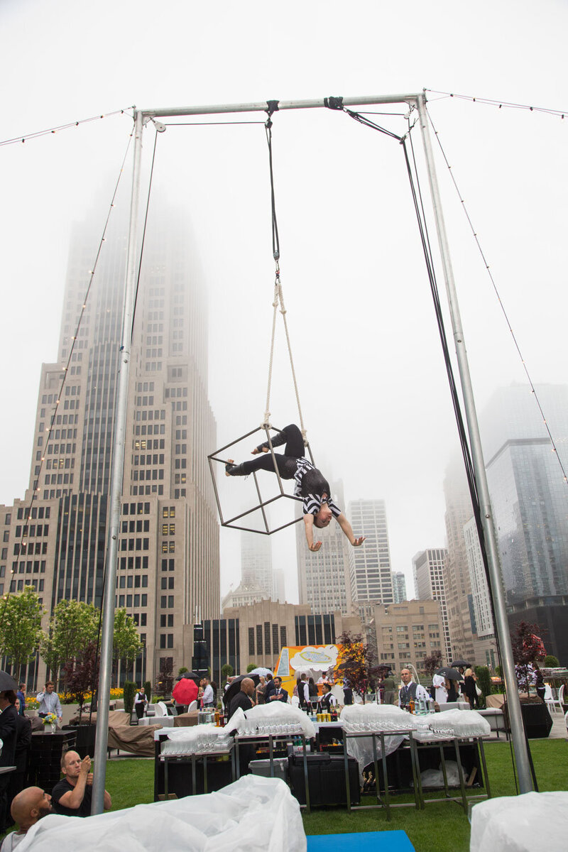 A man is hanging upside down in a cube in front of tall buildings