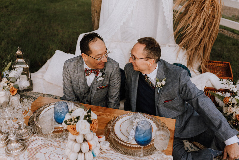 A loving couple in matching plaid suits sits at an elegantly decorated picnic table, gazing into each other's eyes. The outdoor setting features a cozy white canopy, floral arrangements, and vintage tableware.