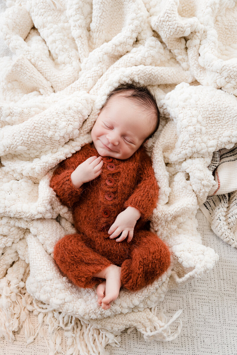 Smiling newborn baby boy in burnt orange fuzzy romper on white textured blankets - professional newborn photography by Glean and Co Boise studio