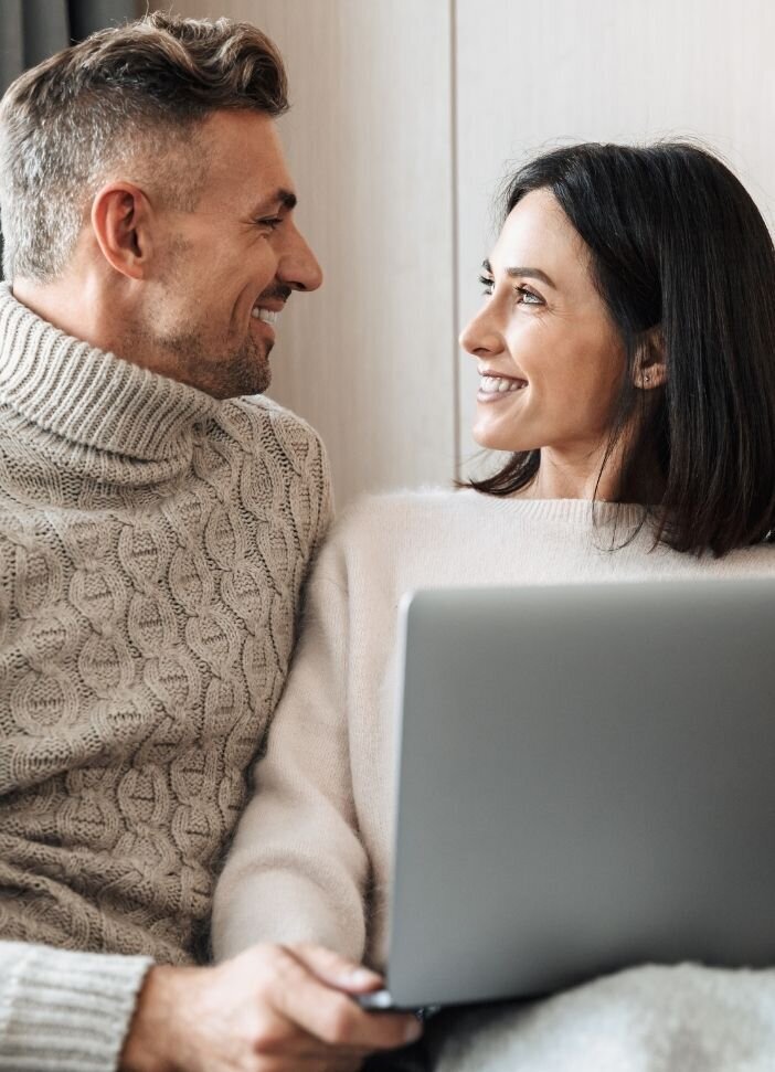 A couple smile at one another while holding a laptop. This could represent finding good results from searching marriage counseling near me. Learn more about our couples assessment, couples therapy program, or other services.