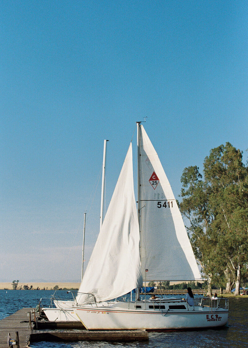 A sailboat with white sails docked on a wooden pier by a calm lake