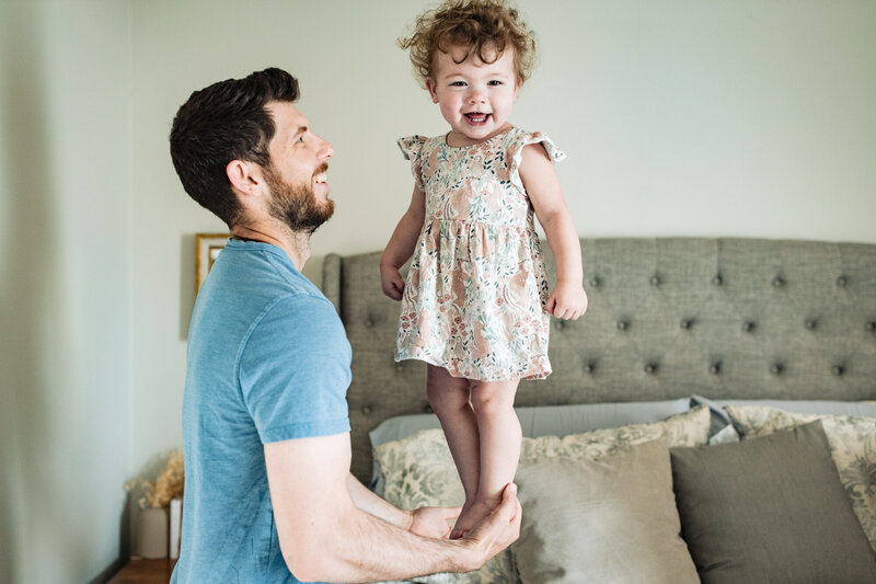 a dad balances his little girl in his hands over the bed while the little girl giggles.
