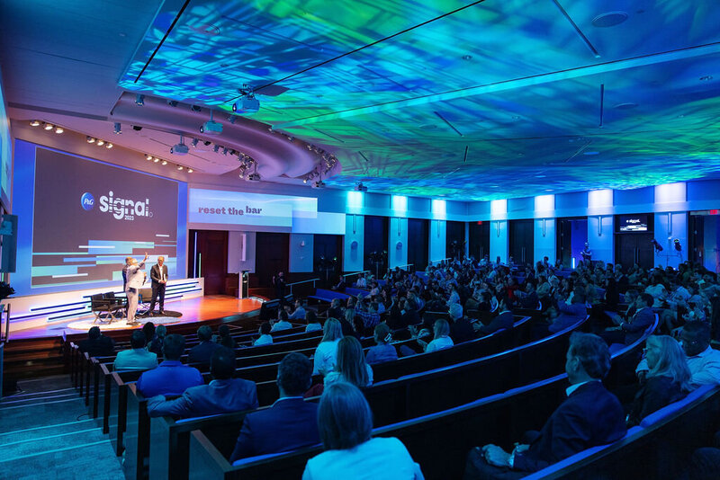 Two speakers on stage at a corporate event with a blue-lit backdrop.