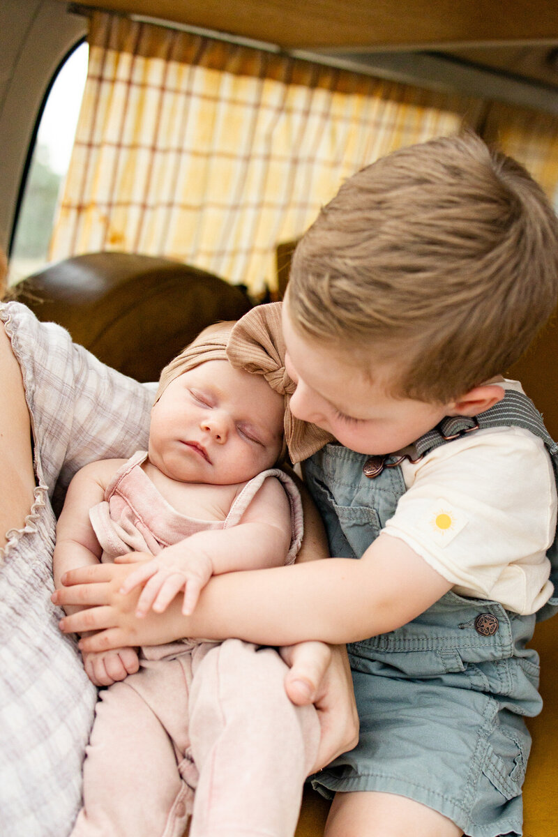 A little boy in green overalls sits in the trunk of a vintage VW bus and hugs his newborn sister.