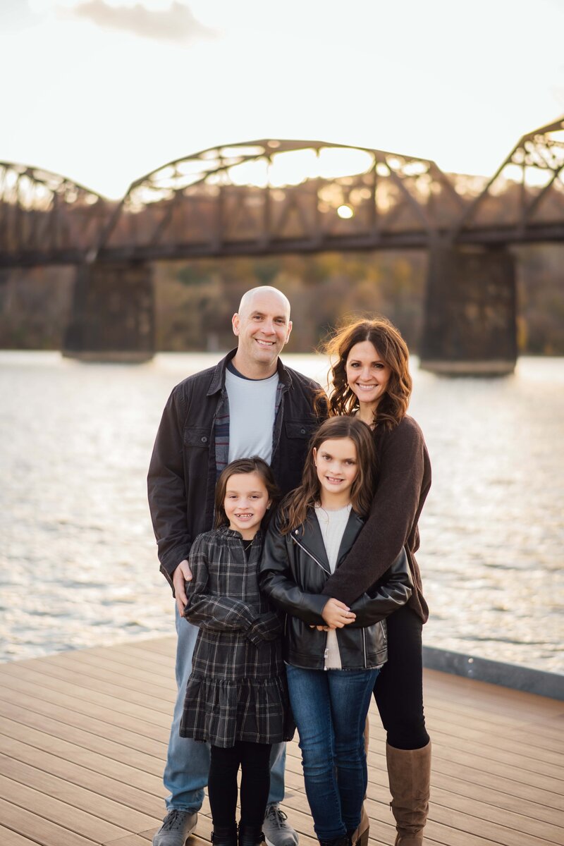 A family of four smiling by a river with a bridge in the background during golden hour, captured by a Pittsburgh photographer.
