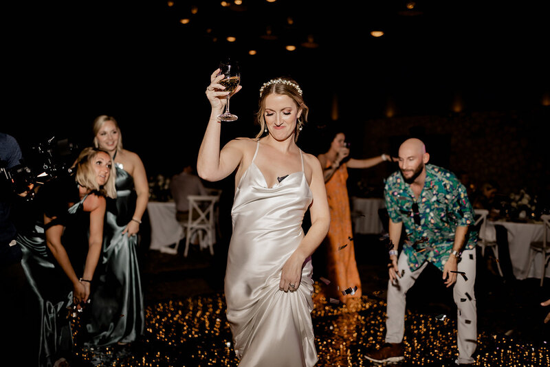A woman in a satin gown holds up a glass of wine while dancing at a wedding reception. Other guests, dressed in formal and festive attire, are also dancing around her on a dimly lit dance floor with scattered lights. The scene captures a lively moment of celebration.