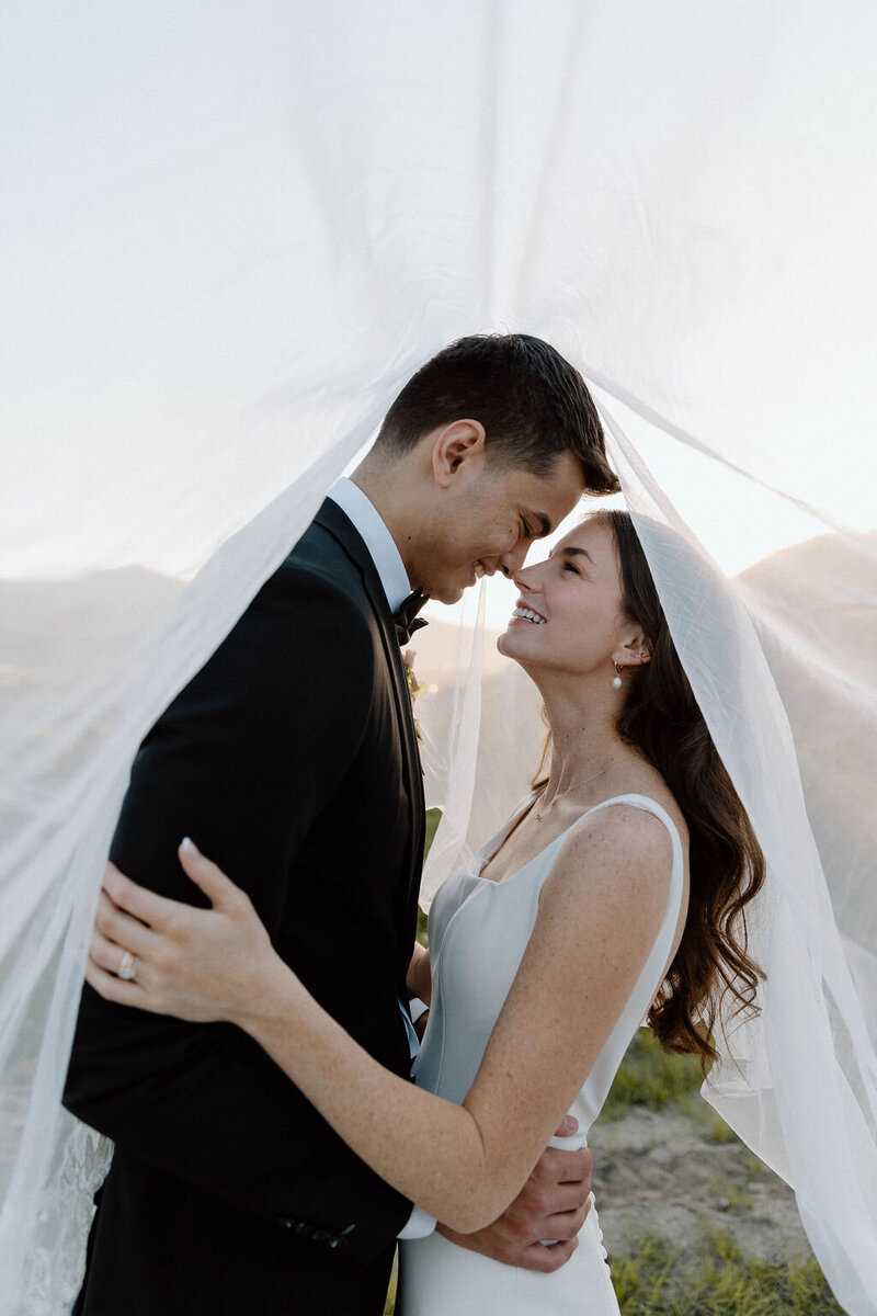 A bride and groom stand nose to nose outdoors, smiling under a sheer white veil, with natural light illuminating their faces.