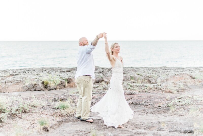 A groom in a white shirt and khaki pants spins his new wife, as if dancing, along the shores of Lake Superior following their northern Minnesota wedding.