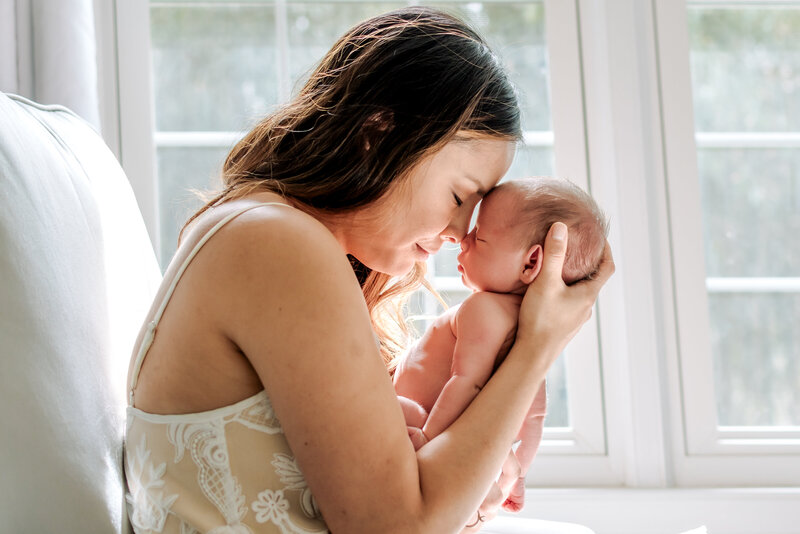 A mom is holding a newborn baby with both of their eyes closed and touching foreheads. They're sitting on a white chair with a window behind them.