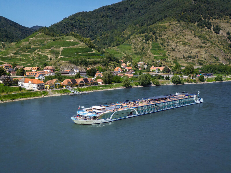 River cruise ship sailing along a wide river with a small village and terraced green hills in the background.