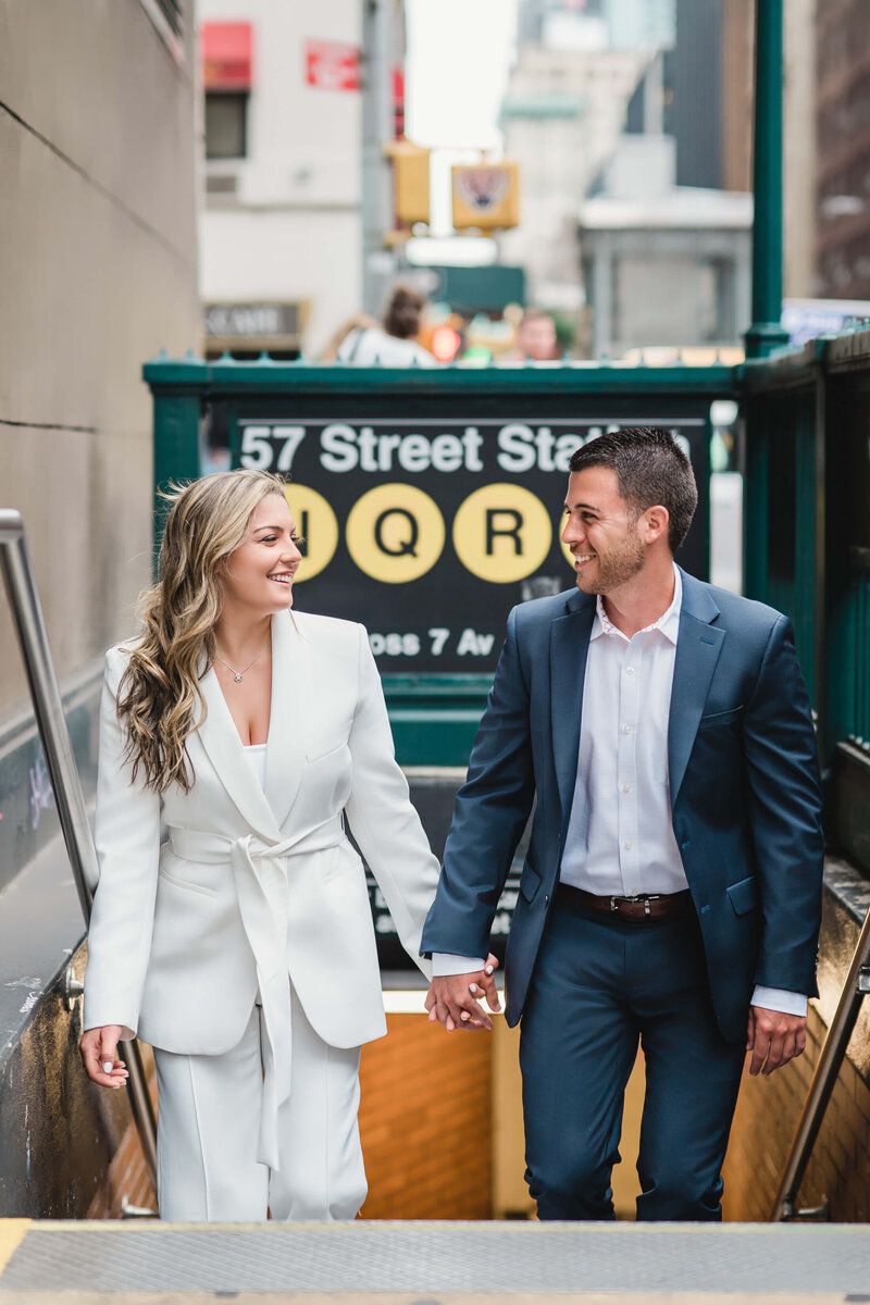 Elegant engagement session: a couple in classic attire emerges from the subway, hand in hand, exchanging smiles as they gaze into each other's eyes
