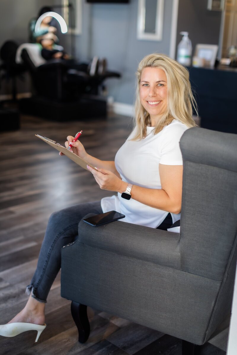 A blonde woman writing on her clipboard smiling in a chair