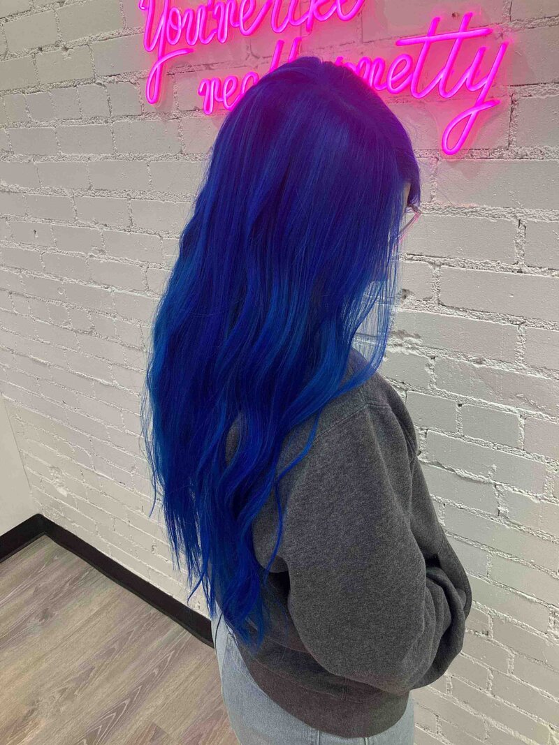 A girl with blue hair standing in  front of a white brick wall and pink neon sign