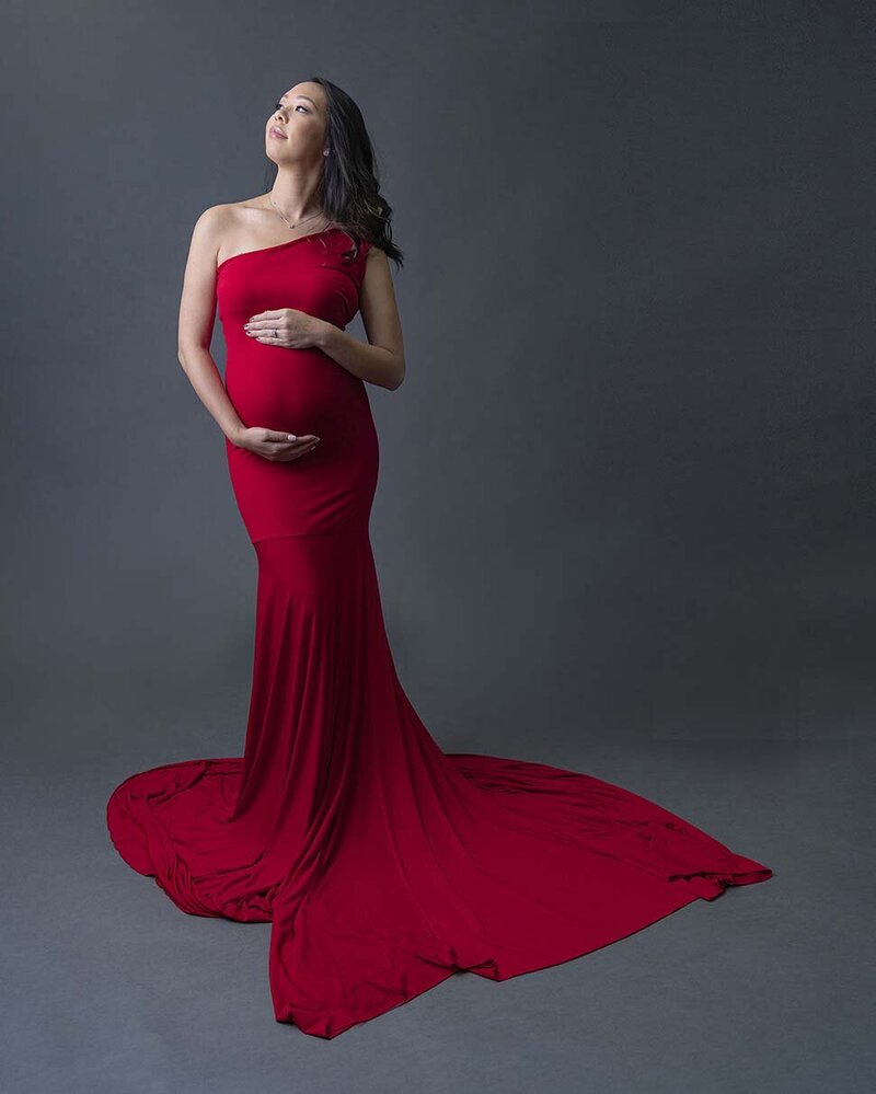 A mother to be stands in a studio smiling up to the light with hands on her bump in a red maternity gown