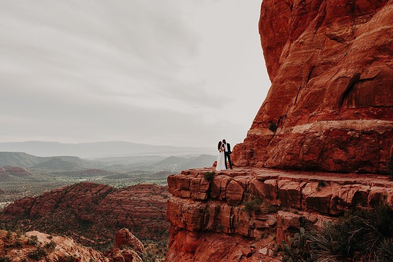 Bride and Groom stand on the edge of a cliff at Cathedral Rock in Sedona taking wedding photos