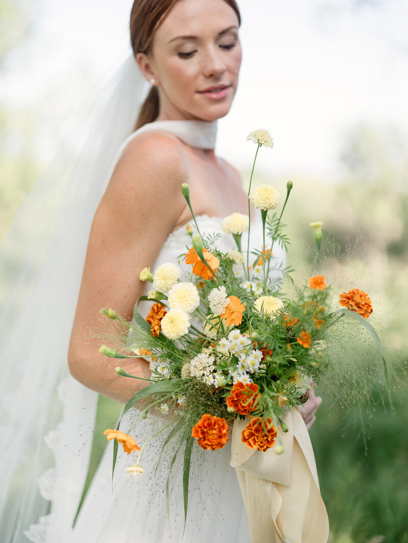 Bride looking down at her wedding bouquet with butter yellow and marigold flowers.