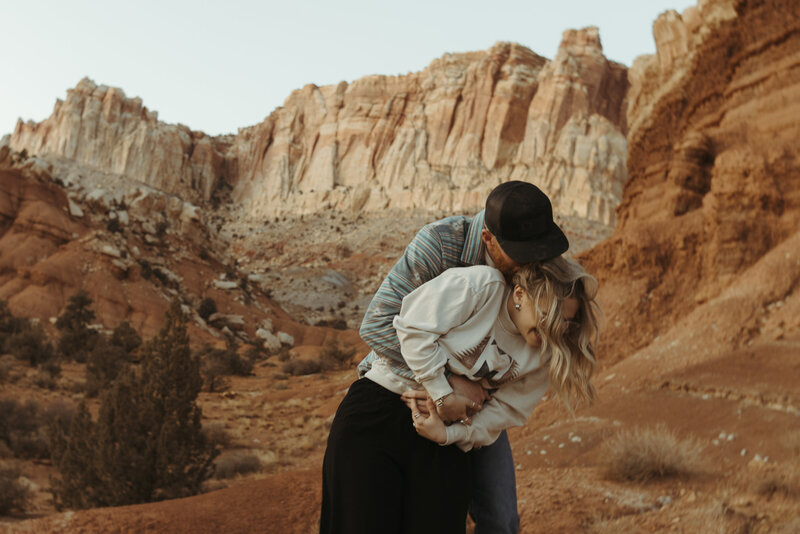 A man and woman dressed in nicer casual clothing holding hands and looking behind them at the beautiful landscape of southern Utah. You cannot see their faces.