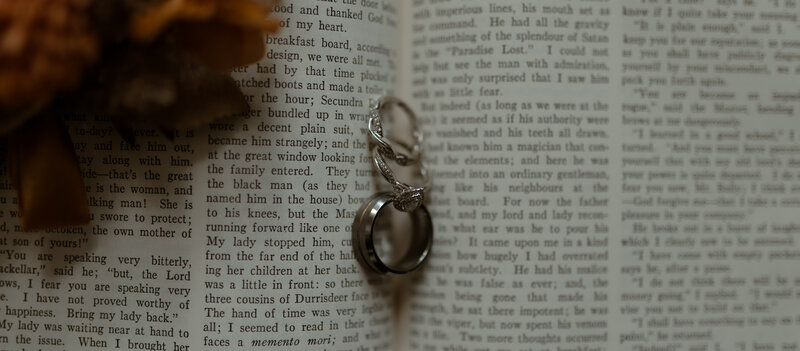 close-up shot of two intertwined wedding rings resting on the pages of an open vintage book. The engagement ring features a delicate diamond set in a silver band, while the groom’s ring is a sleek, polished band. 