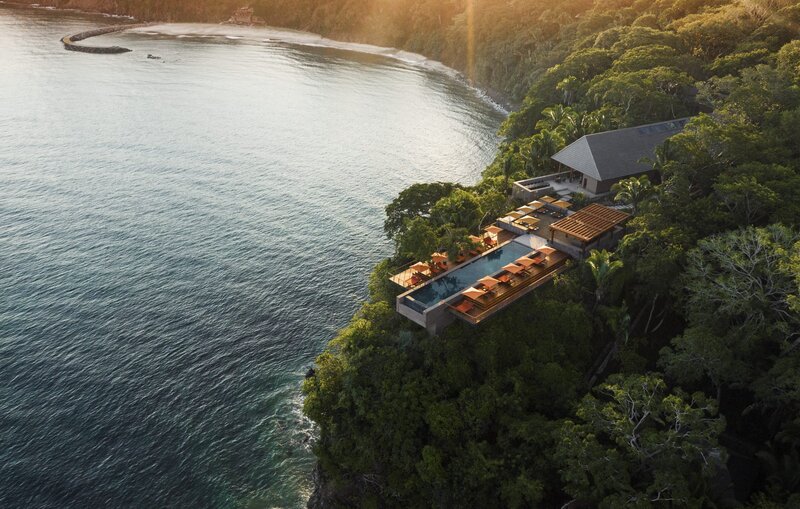 A breathtaking aerial view of a secluded luxury resort perched on a lush cliffside overlooking crystal-clear waters at sunset. Experience bespoke private island escapes, exclusive resort stays, and high-touch travel planning with Nadia Forte Travel & Events.