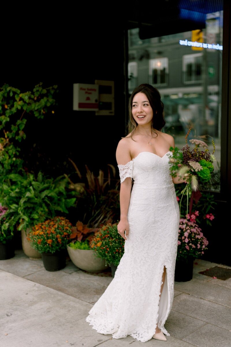 A smiling woman in an off-the-shoulder white dress holding a bouquet, standing on a city sidewalk.