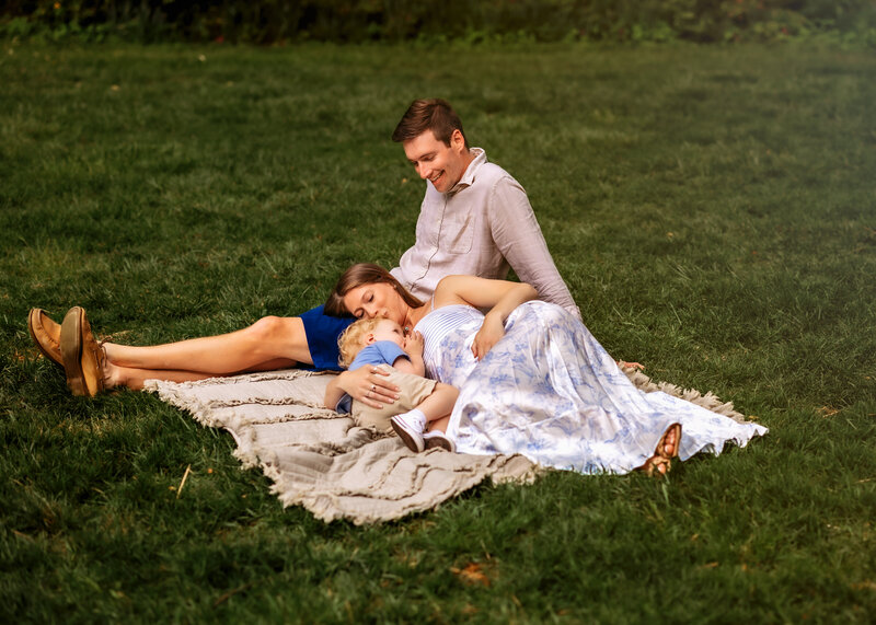Dad sits on blanket with mom laying on his land with hands on her pregnant bump and cuddling two year old snuggling close. 