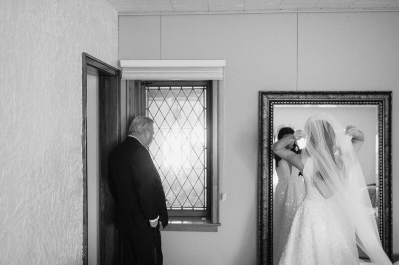 Black and white photo of the father of the bride looking out a window while the bride fixes her veil. Photo by Anna Brace an NE Wedding Photographers.