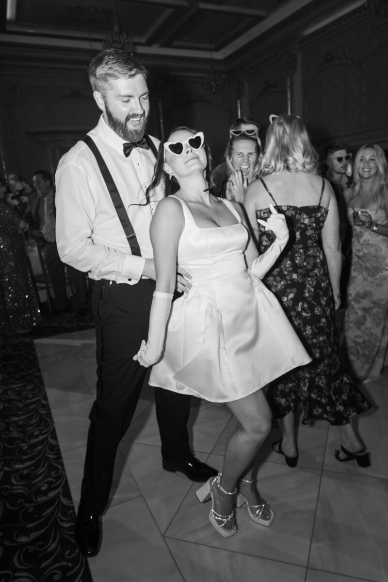 Bride wearing heart sunglasses, gloves and a short party dress dancing with her husband during their wedding reception at Brigalias