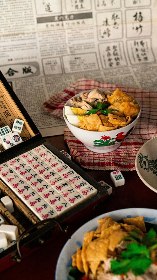 A bowl of chicken soup garnished with egg and greens sits on a checkered towel. A mahjong set lies open nearby, with tiles scattered, atop a surface with Chinese newspaper pages. The scene is cozy and nostalgic.