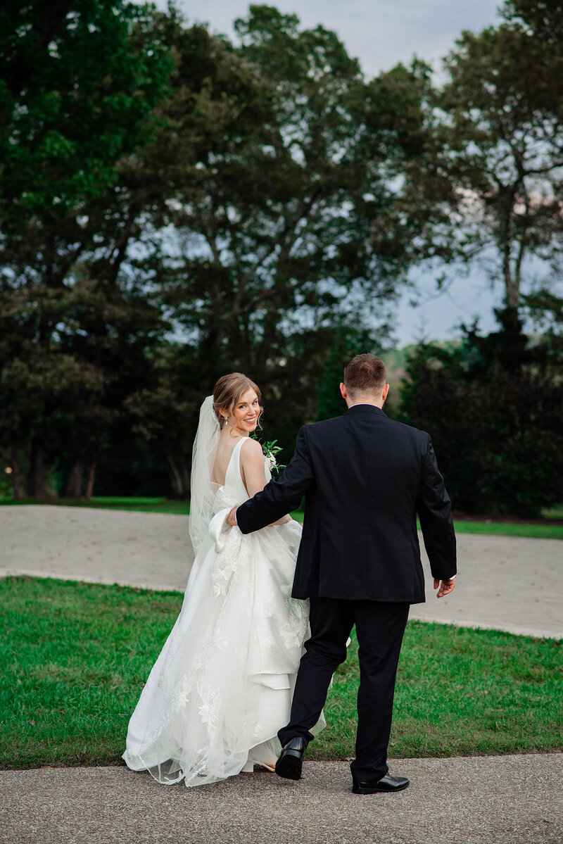 Groom carrying brides train and she is looking over her shoulder smiling at the camera