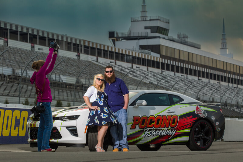 Behind-the-scenes photo on the track at Pocono Raceway during an engagement session, featuring a couple with the dramatic backdrop of the iconic racetrack, capturing unique and dynamic moments.