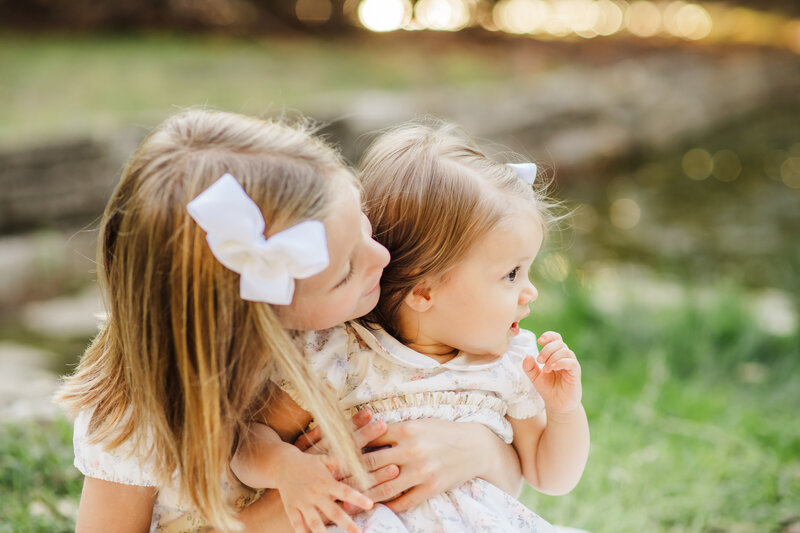 Two young girls are sitting together on grass at Mills Pond Park in Austin, TX, captured by an Austin Texas Photographer.