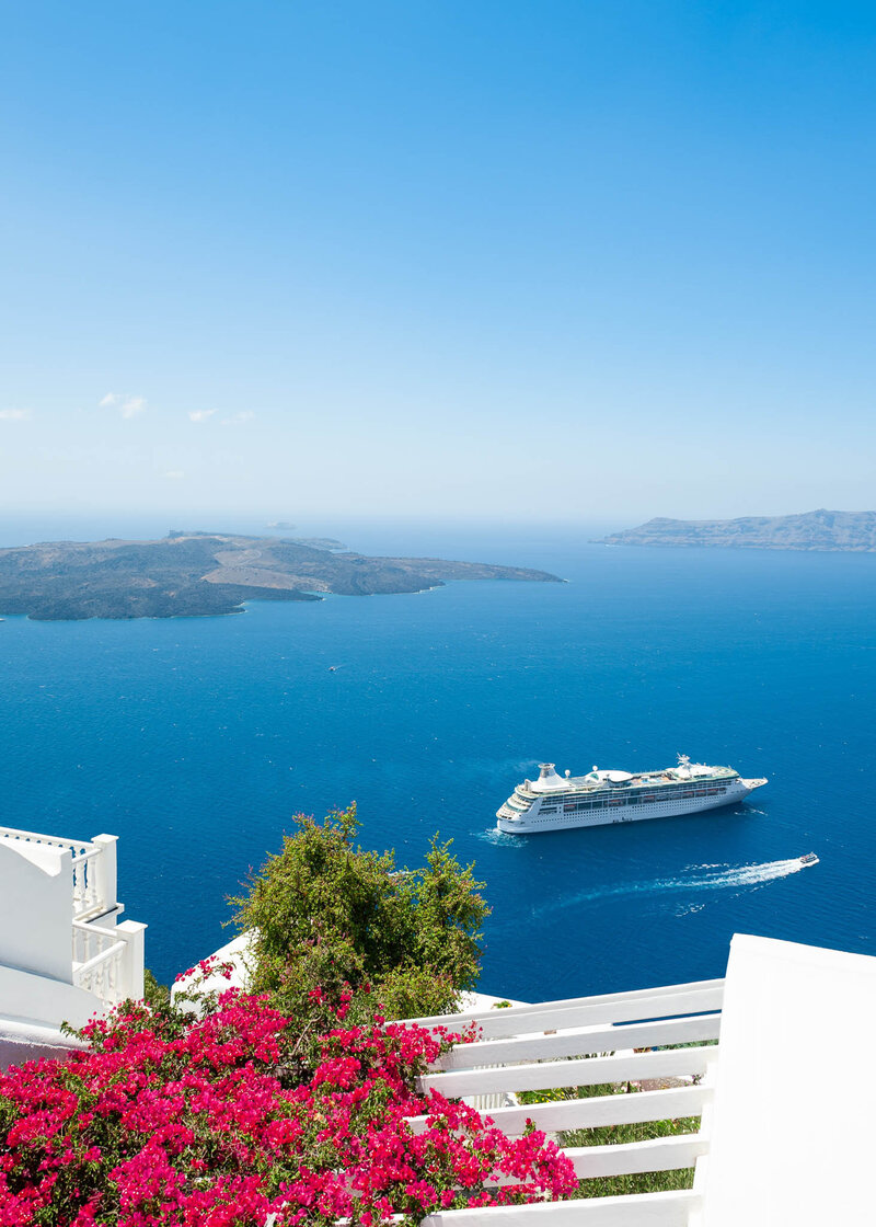 Cruise ship sailing on blue sea near white buildings and pink flowers on a sunny day in Santorini, Greece.