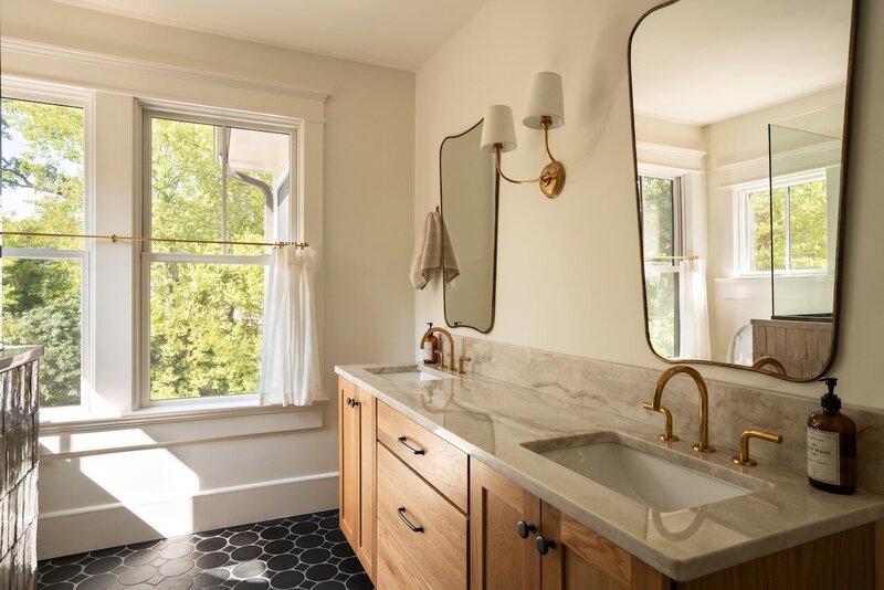 Master bathroom double vanity with wooden cabinets, organic metal-rimmed mirrors, double wall sconces, backyard window, black tile floor, and brushed gold faucets.