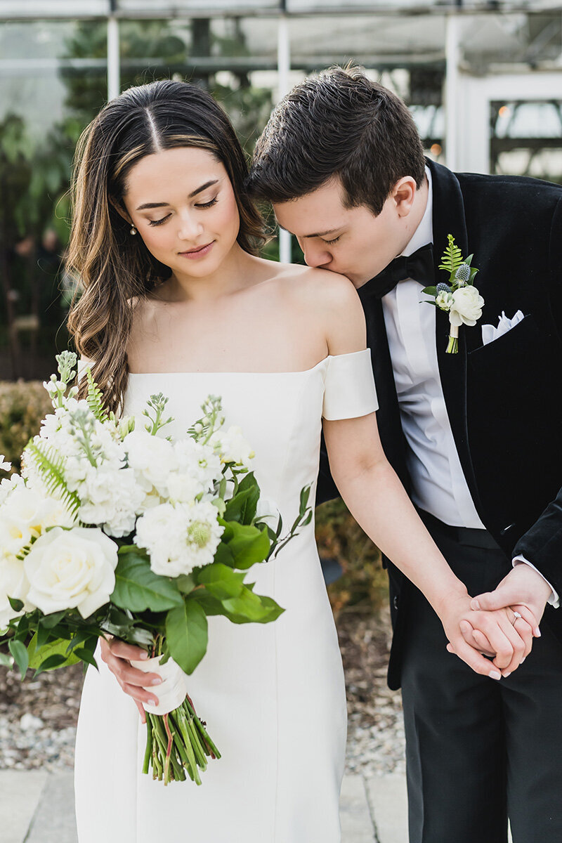 Bride and groom share a romantic first kiss under the greenhouse canopy at Planterra Conservatory, surrounded by lush greenery, string lights, and emotional guests during a black tie wedding ceremony.