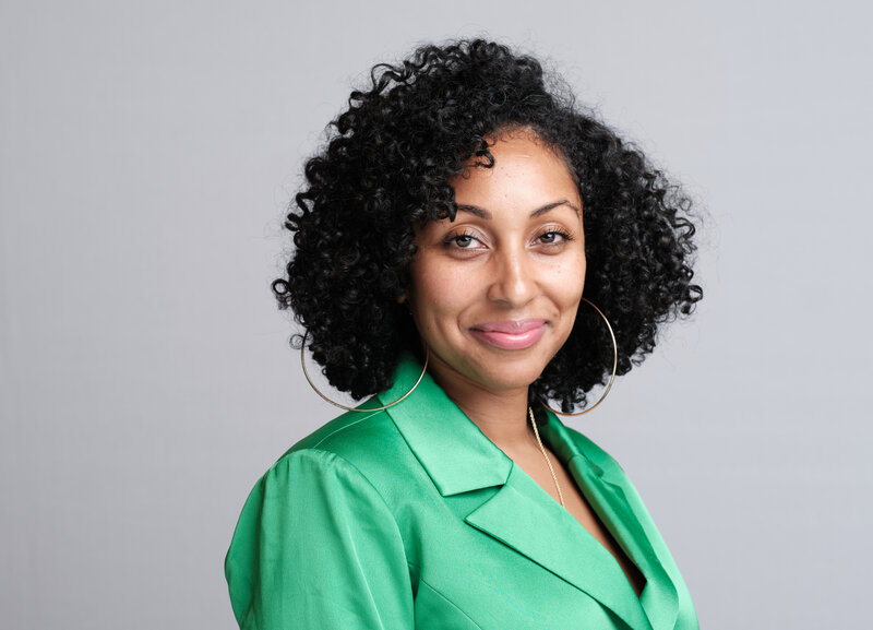 A woman in a black suit and purple blouse poses for a photo in front of a wood panel.