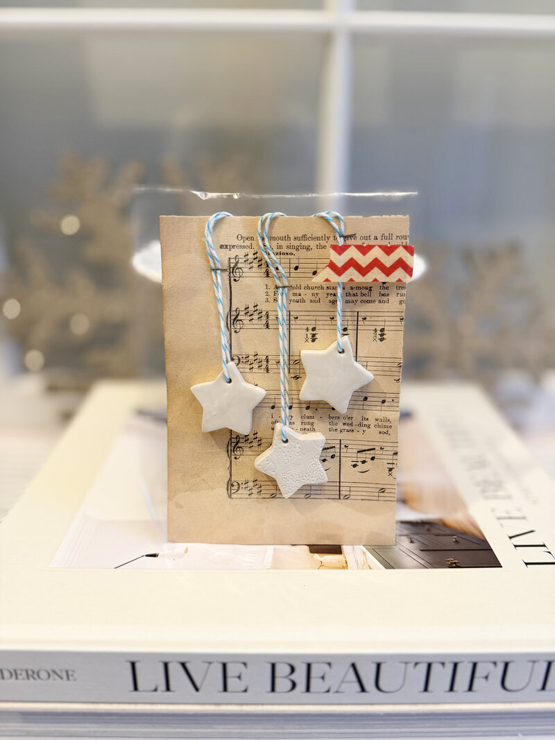 Set of three petite star-shaped clay ornaments from Sweet Pea Ink Creative, each tied with blue and white baker’s twine and displayed against vintage sheet music. Packaged with red chevron tape on a holiday-themed backing card.