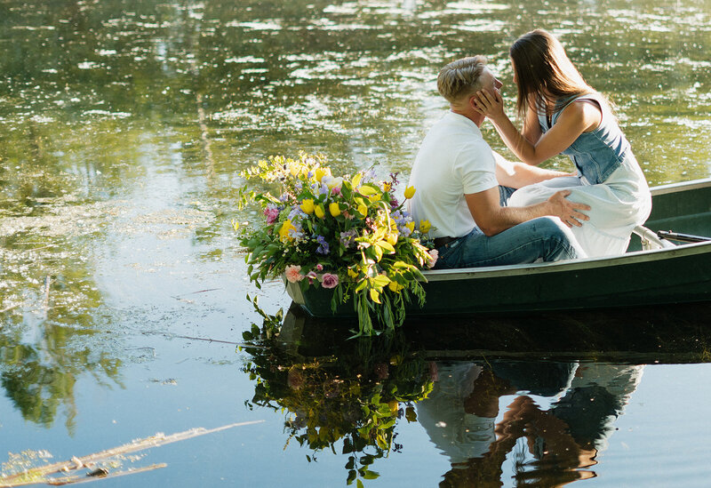 Couples photography adventure session in Rexburg Idaho. A woman is kissing her boyfriend or fiance in a rowboat in a pond. At the front of the rowboat is a large bouquet of flowers.