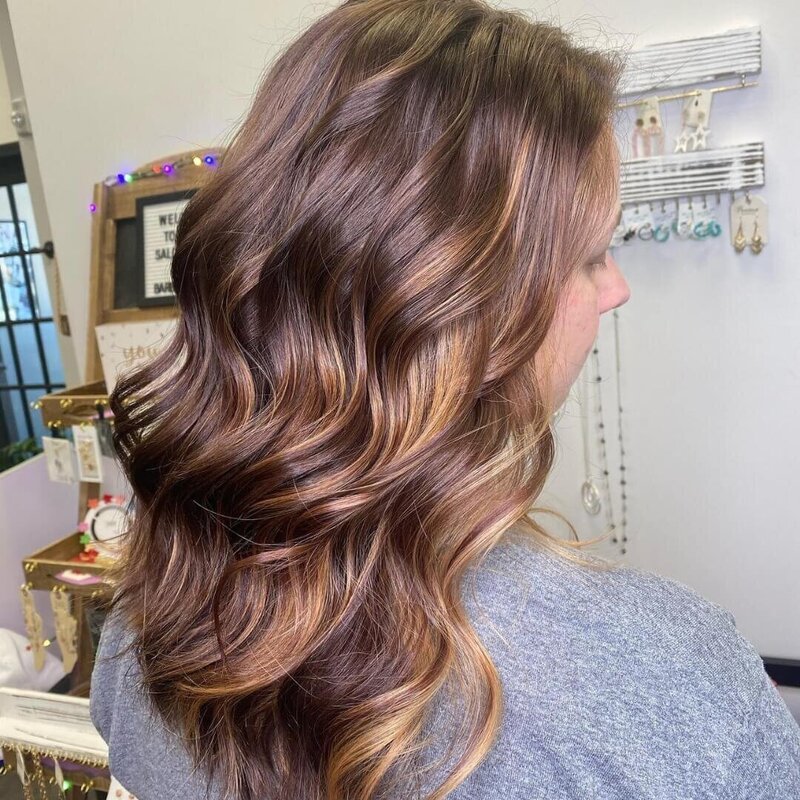 A woman with long, shiny brunette waves and subtle highlights, showcasing a beautifully styled look created by 212 Salon, Spa, & Barbershop.