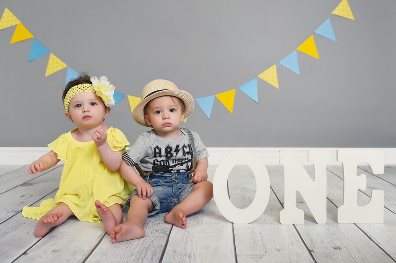 Twin babies celebrating their first birthday; the girl in a bright yellow dress with a flower headband and the boy wearing a straw hat and suspenders, sitting together with "ONE" letters and yellow and blue bunting decorations.